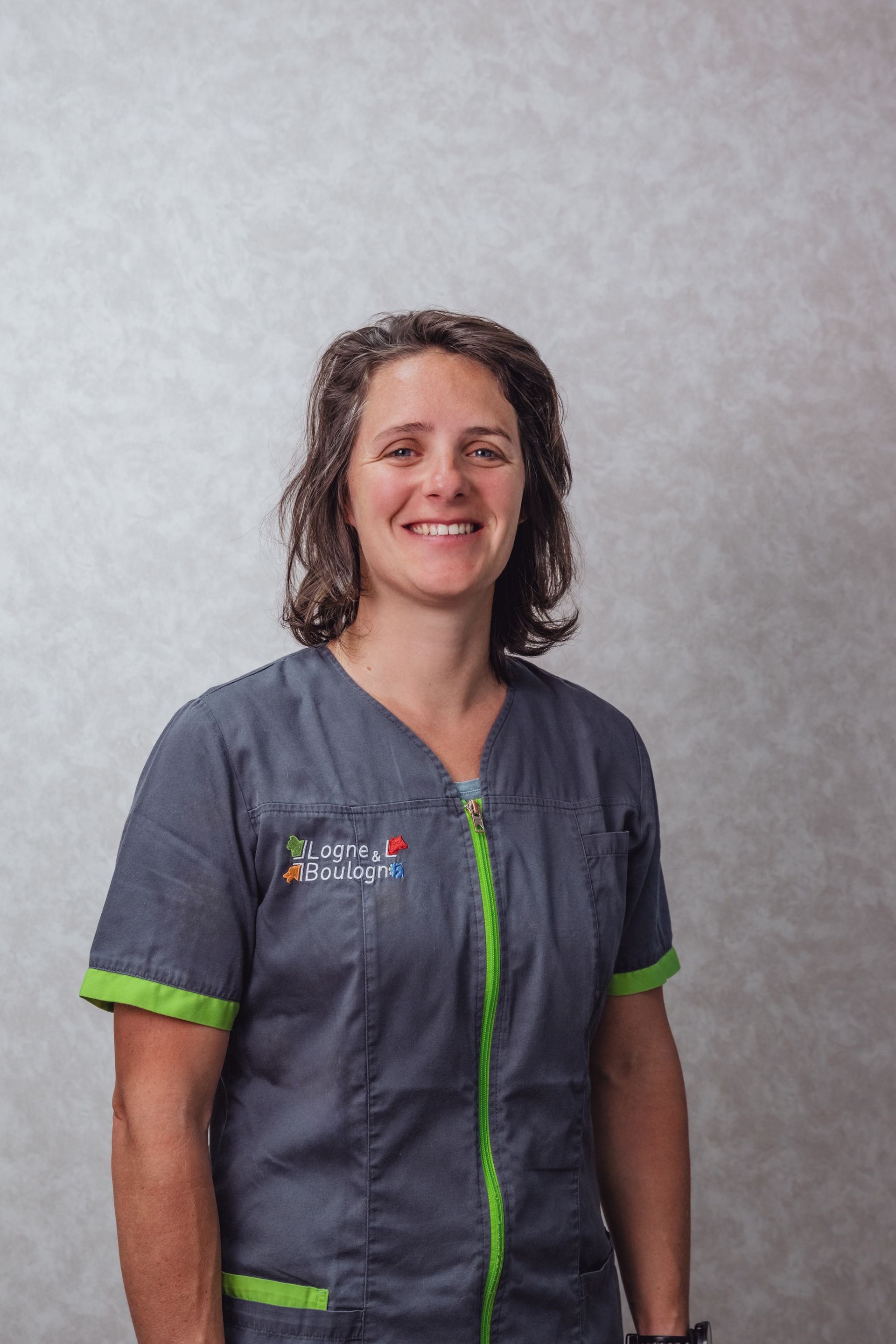 Une femme en blouse grise sourit, debout devant un mur texturé.