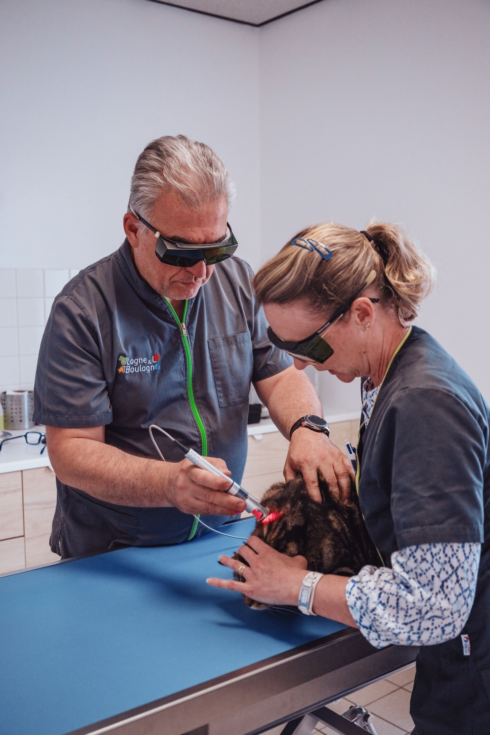 Deux personnes portant des lunettes de sécurité utilisent un laser sur la tête d'une personne allongée sur une table bleue. Contexte clinique.