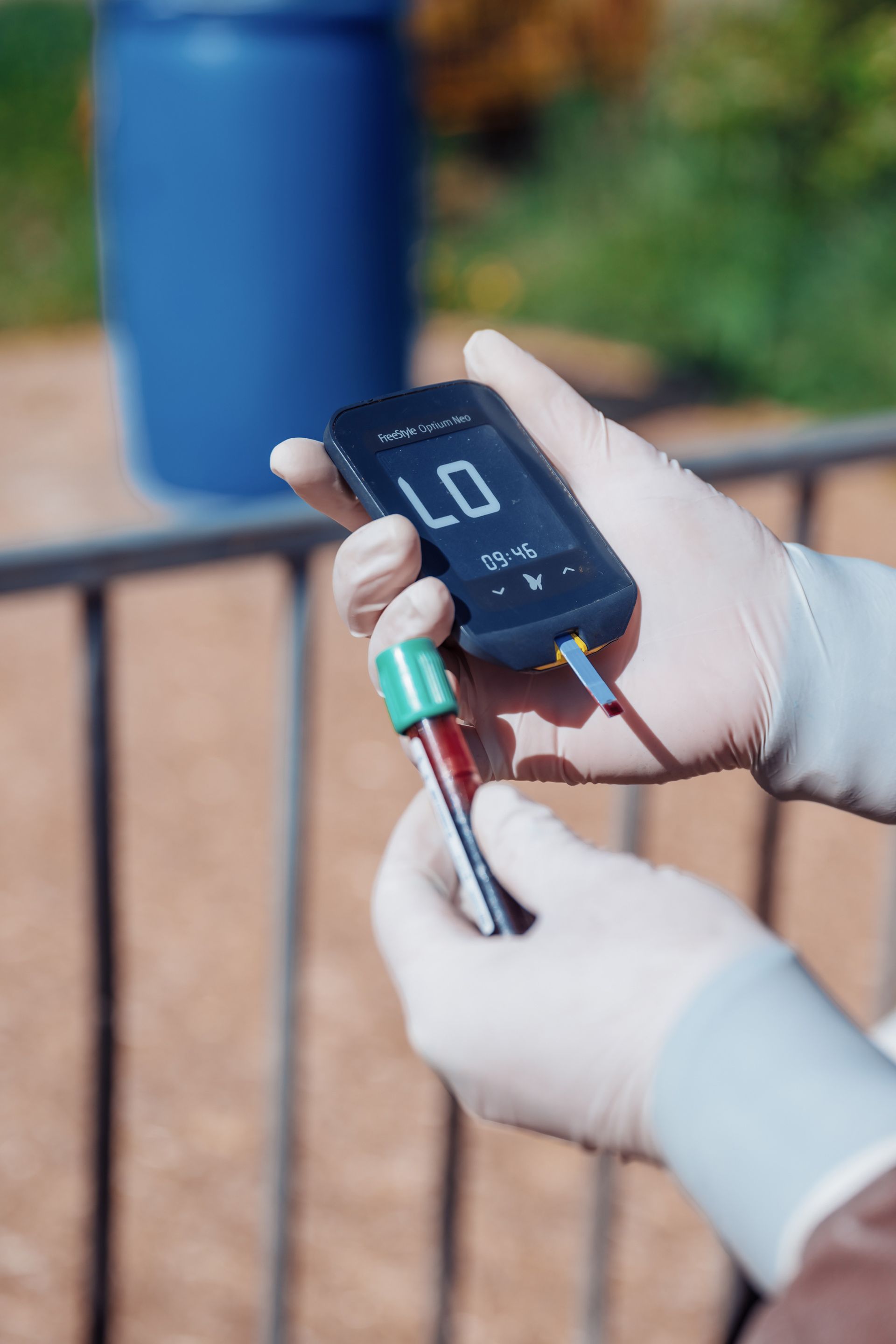 Des mains gantées tenant un glucomètre affichant « LO » et un flacon de sang à l'extérieur, à côté d'un baril bleu.