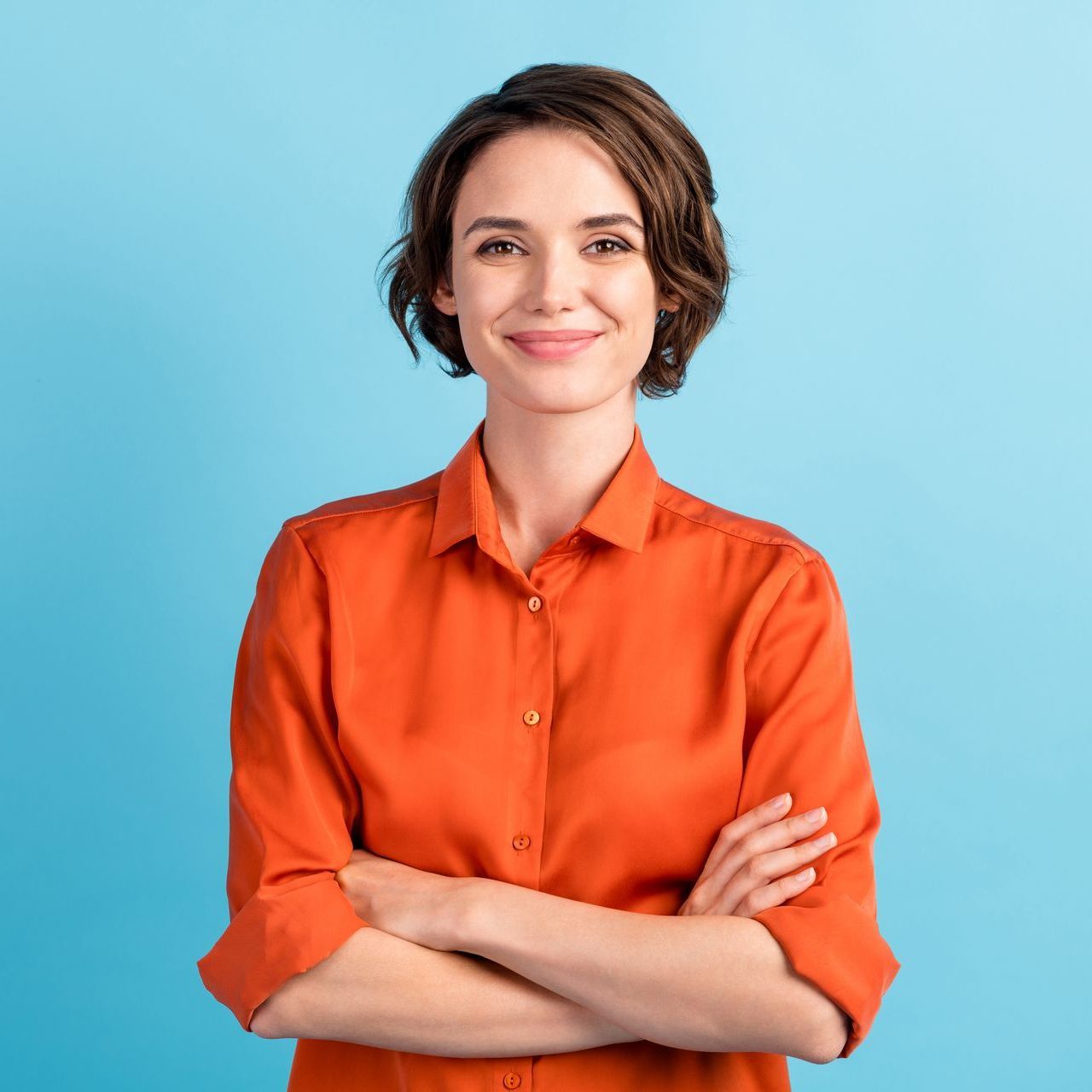 A woman in an orange shirt is standing with her arms crossed and smiling.