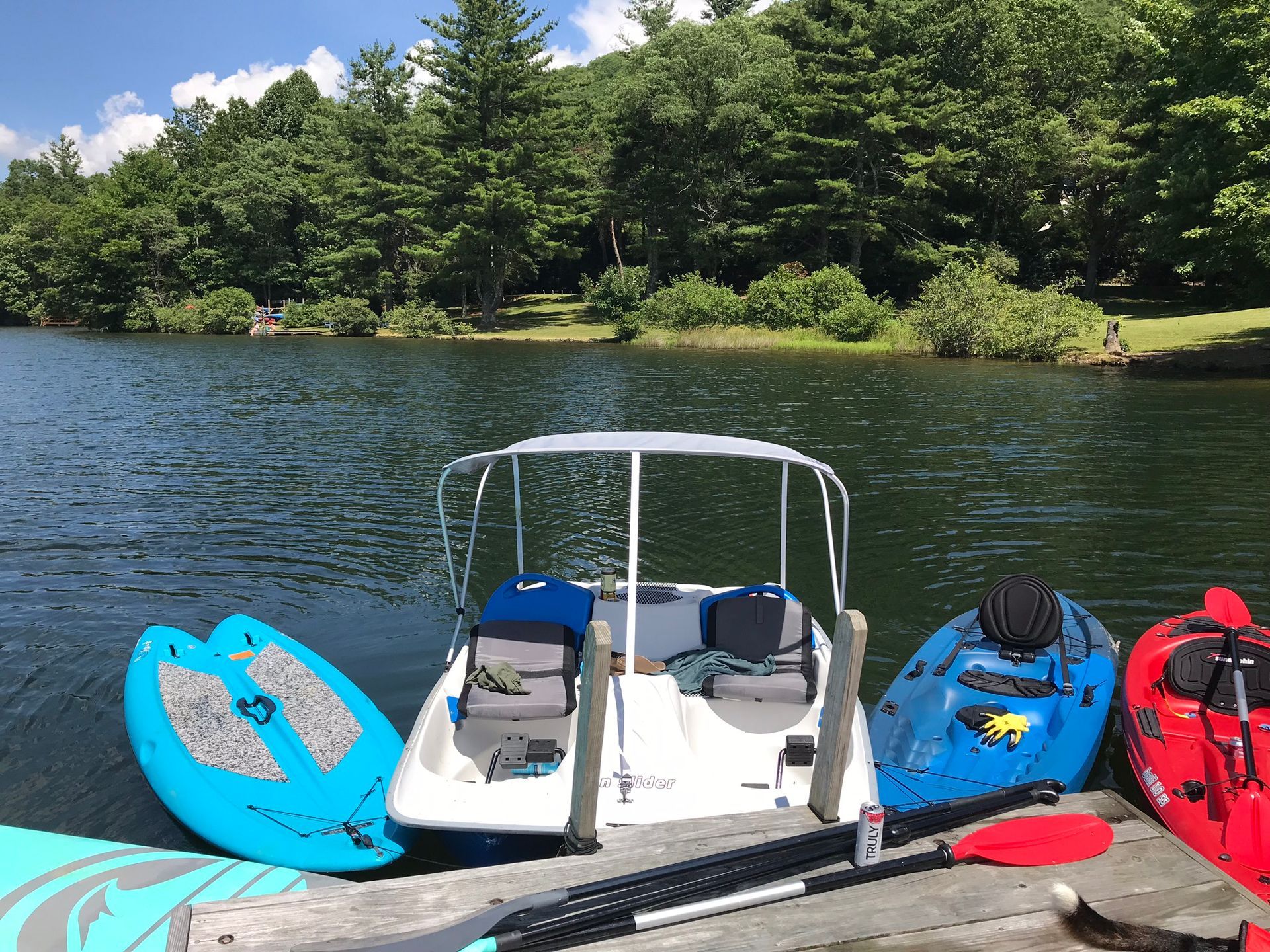 Boat docked with paddle boards and kayaks on a lake. Trees in background, sunny day.