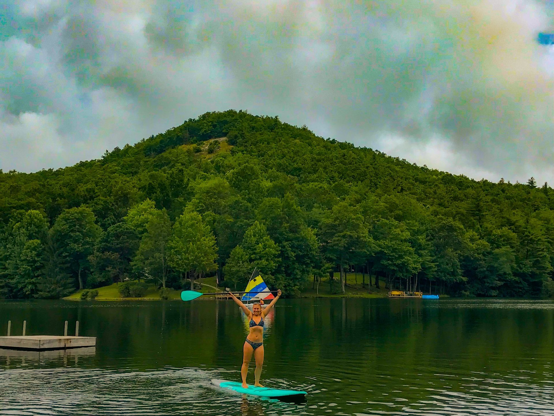 Woman on paddleboard, arms raised, on a lake, mountain in background under cloudy sky.