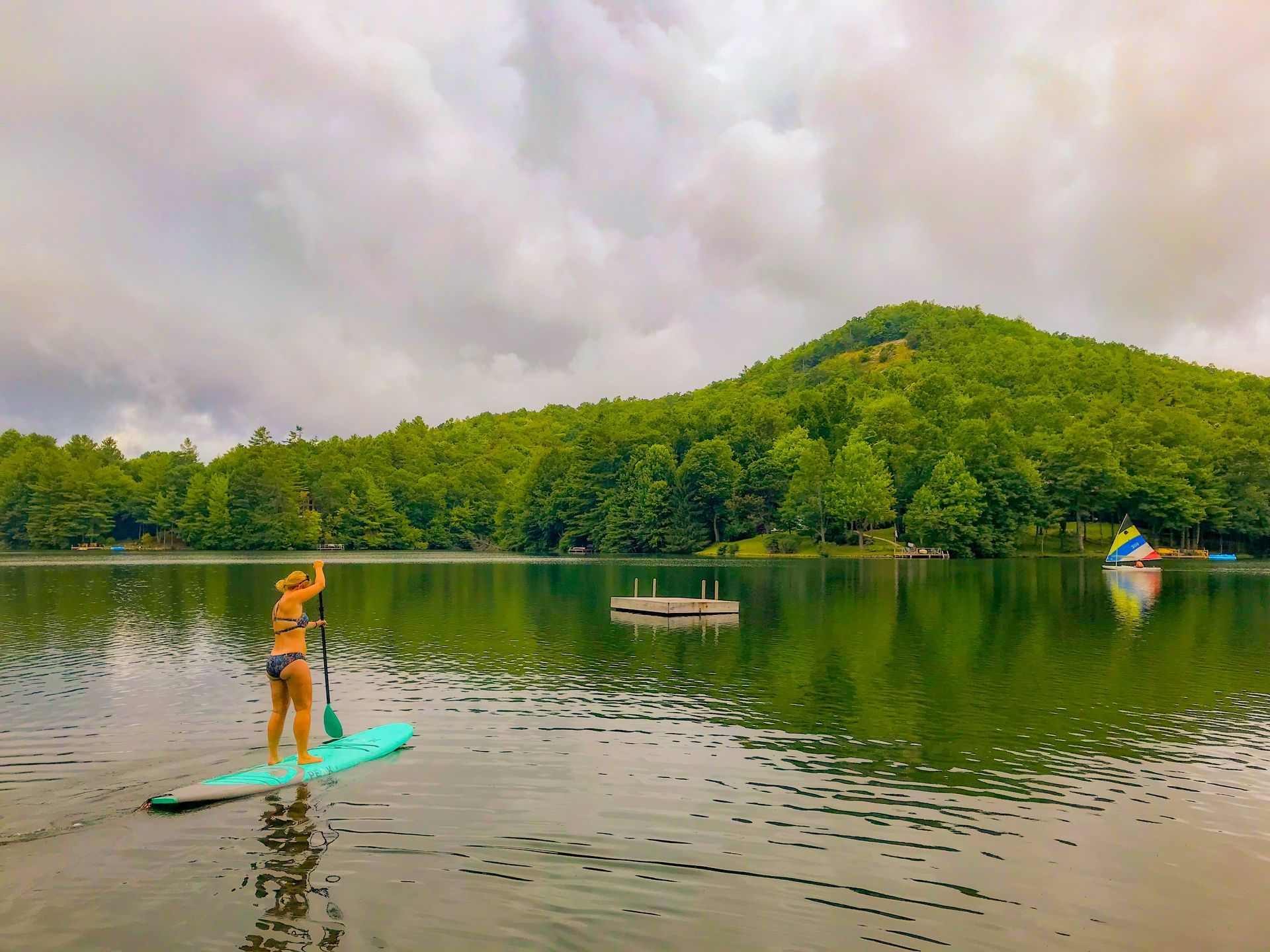 Woman paddleboards on a lake under cloudy sky, surrounded by green trees.