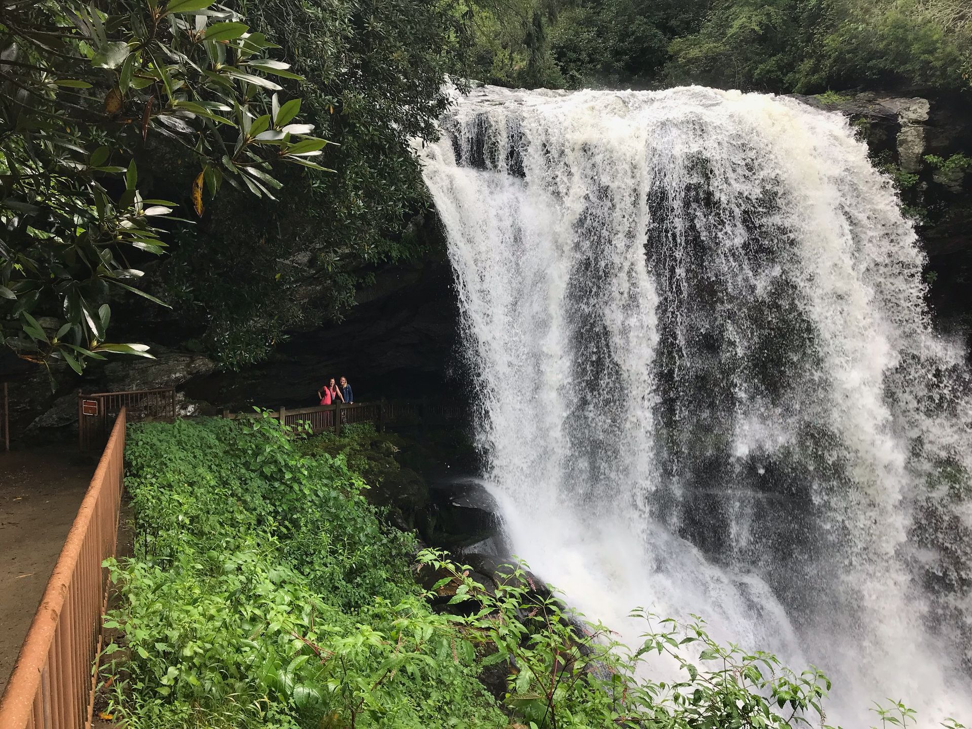 Waterfall cascading over rocks; path with railing and people viewing. Green foliage surrounds.