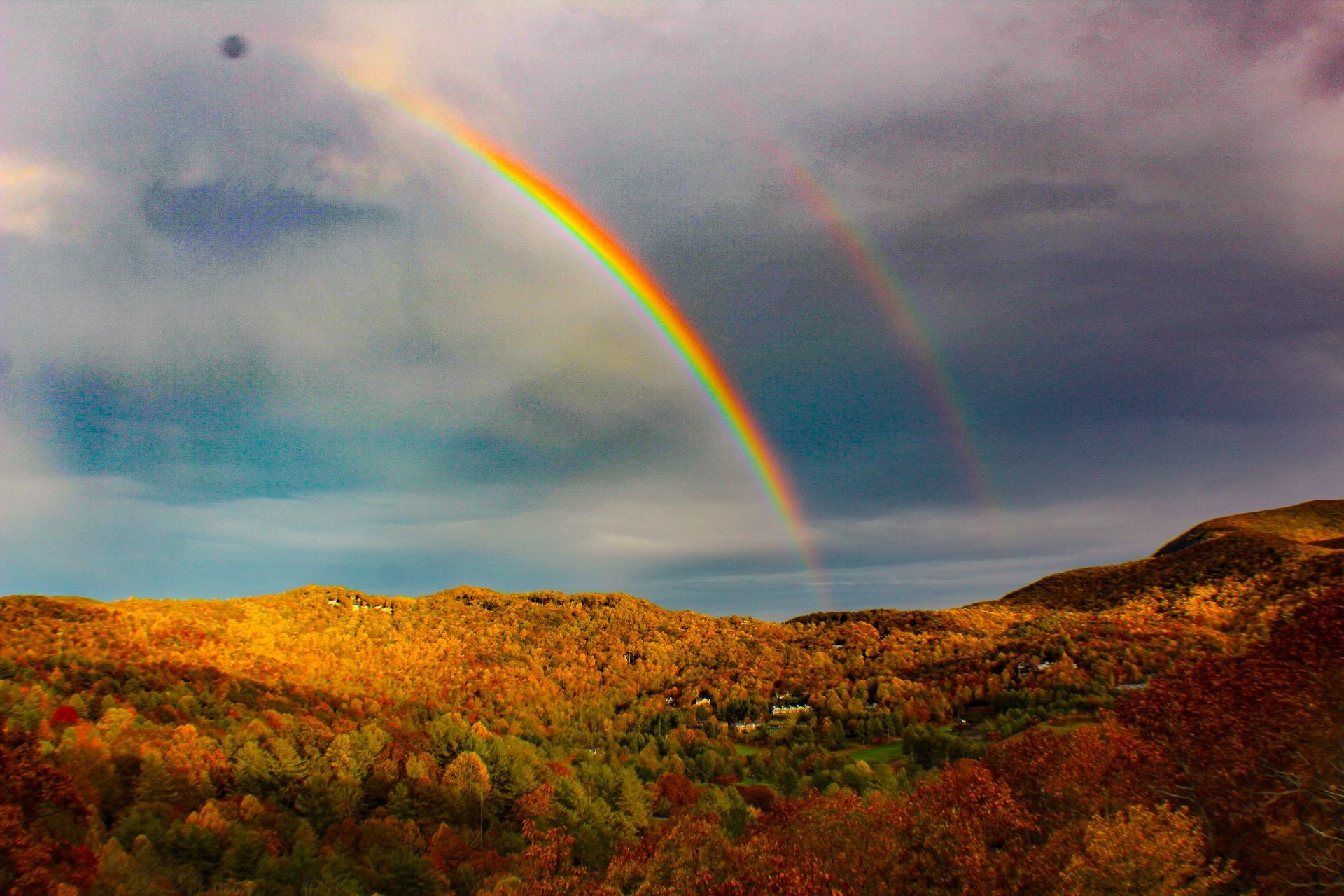 Double rainbow arches over a forested mountain landscape, lit by golden sunlight.