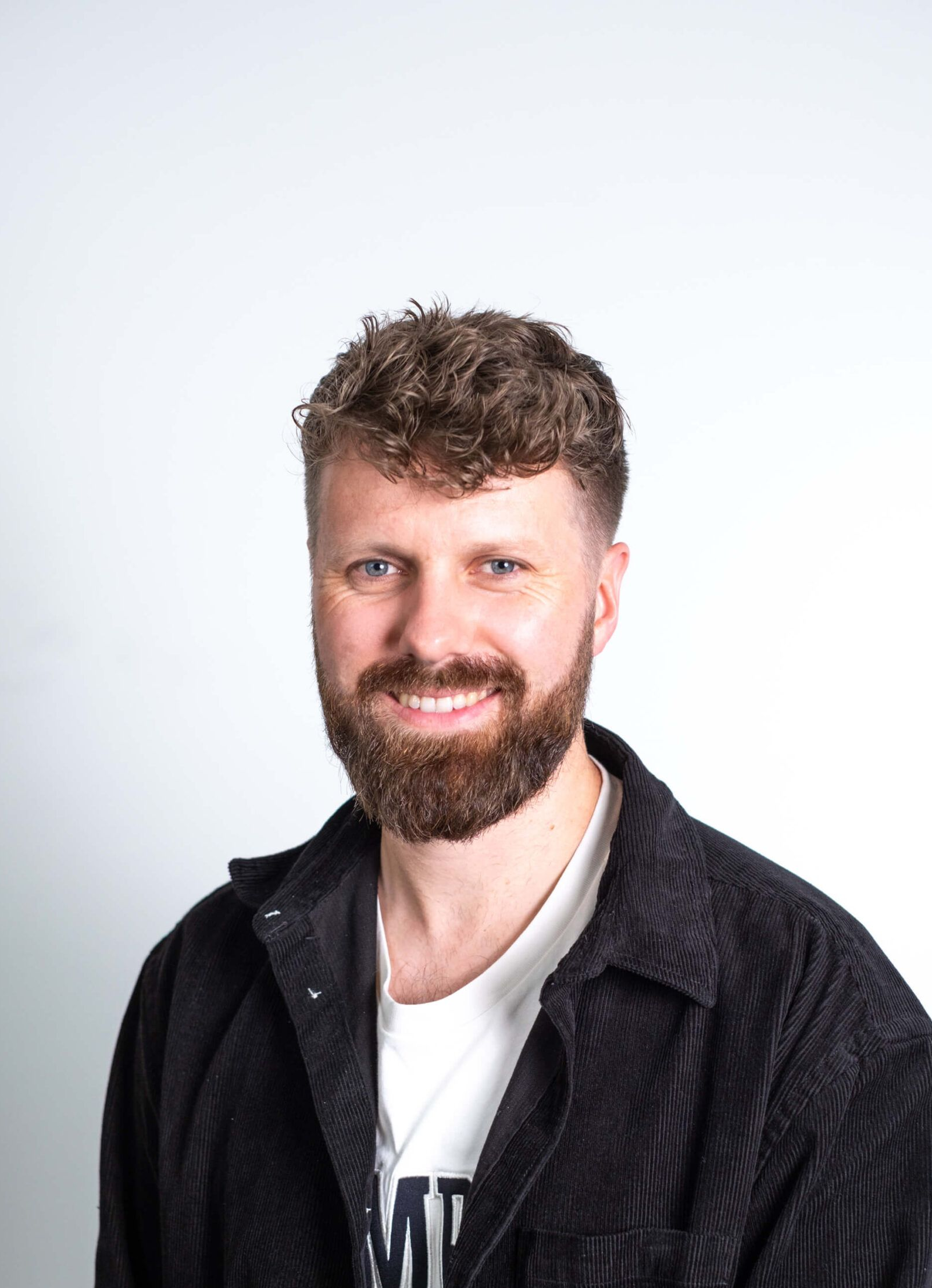 Man with brown curly hair and beard smiling, wearing black corduroy jacket and white shirt.