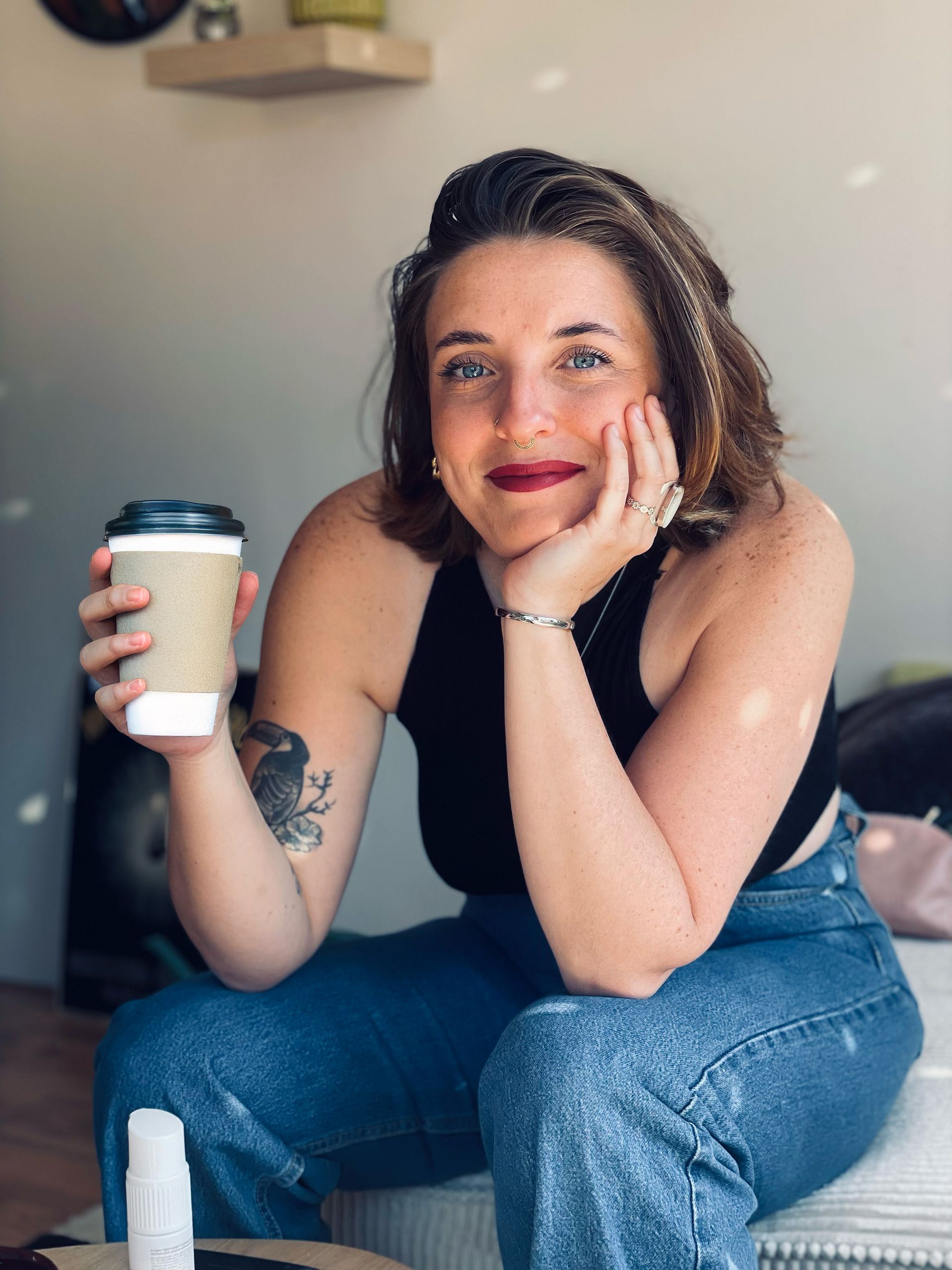 Woman in black top and jeans holding coffee, smiling, resting chin on hand.
