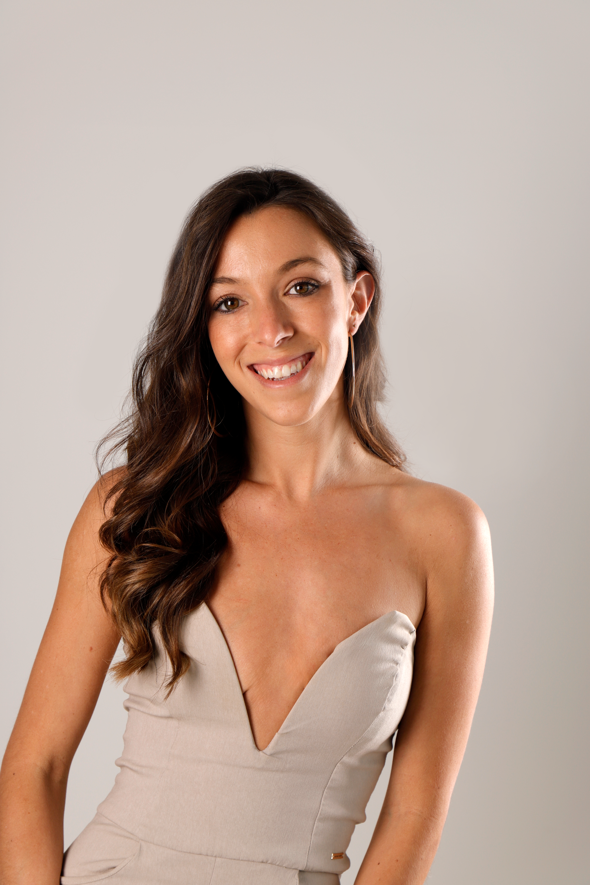 Woman in a strapless beige dress smiles, standing in front of a neutral backdrop.