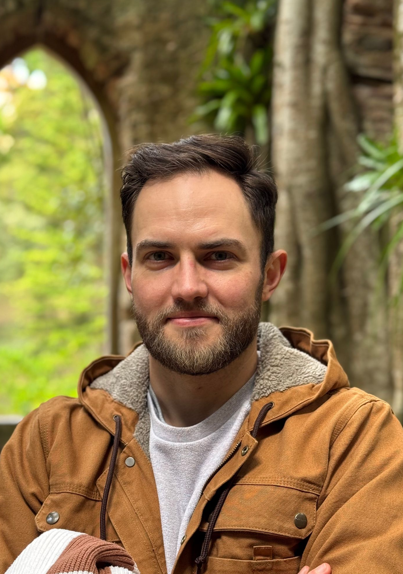 Woman with slicked-back hair and hoop earrings, posing with an orange wall background, wearing a tan and white shirt.