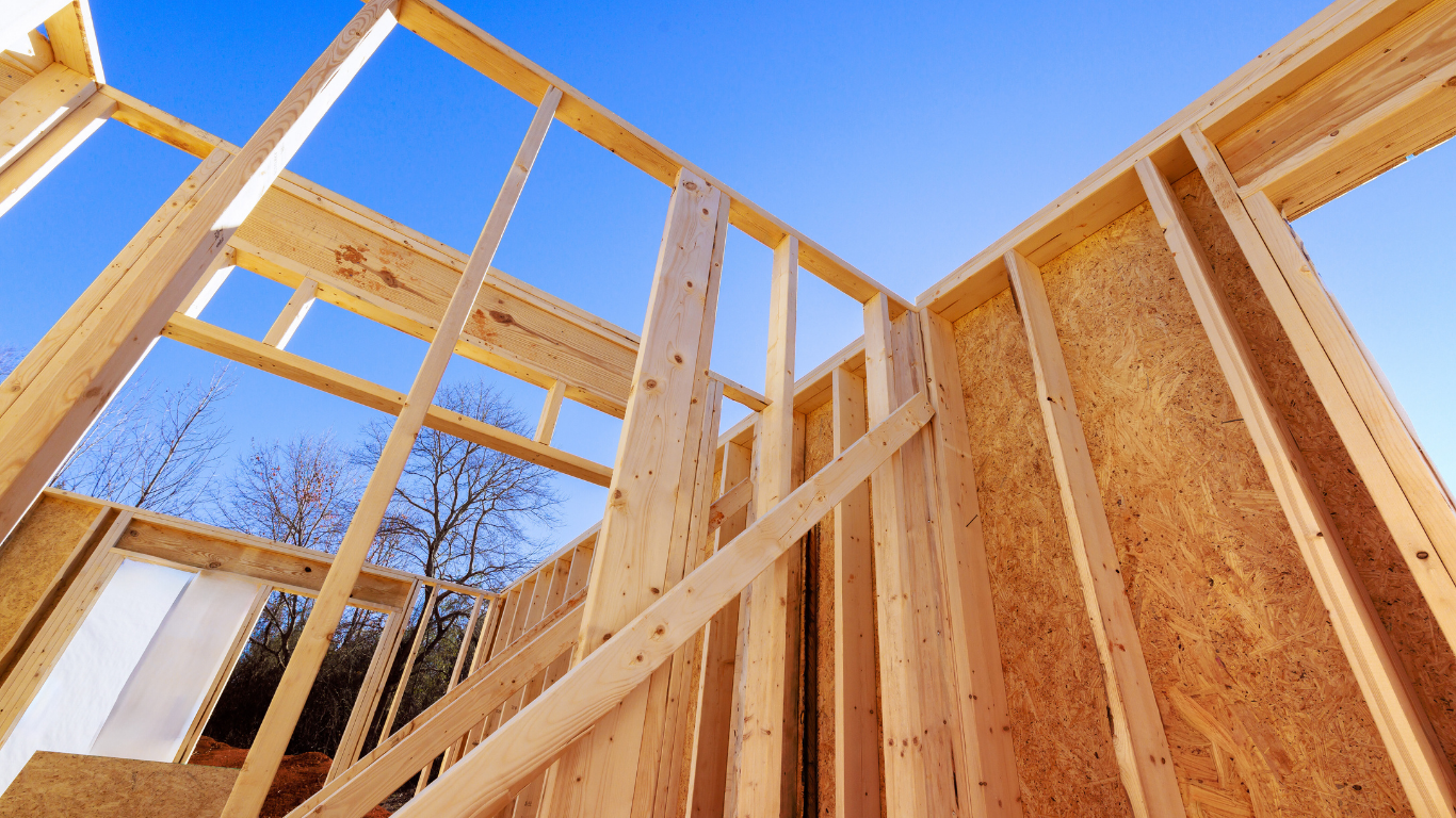 Wooden house frame against a blue sky, under construction, showing beams and wall supports.