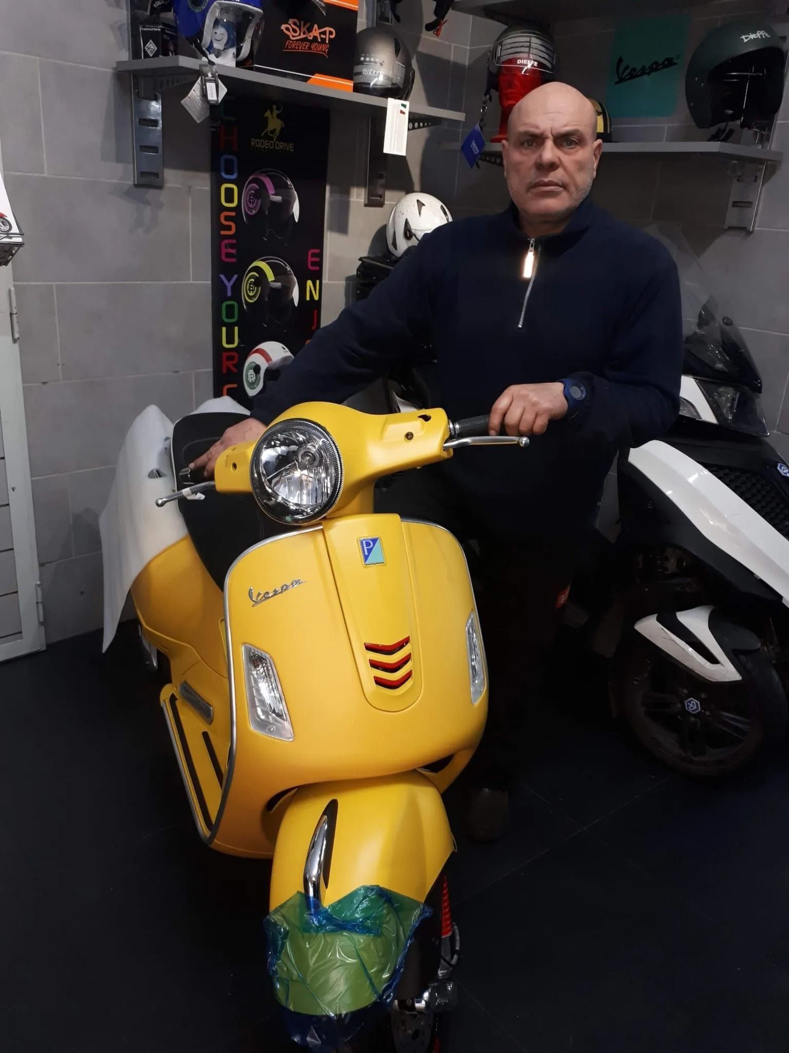 Uomo in posa con uno scooter giallo in un negozio, sullo sfondo altri scooter e caschi.