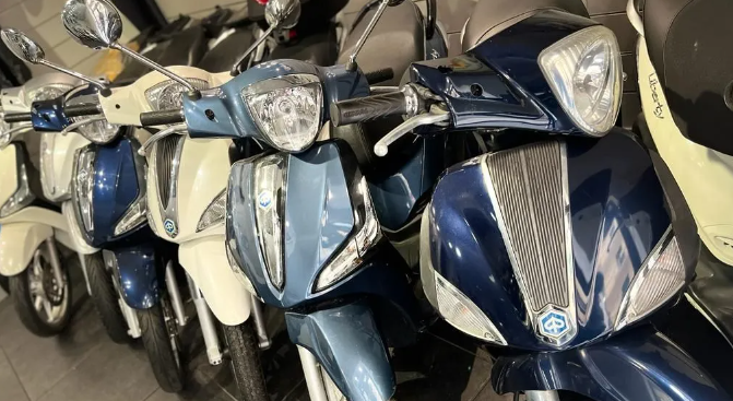 Fila di scooter blu e bianchi parcheggiati al chiuso, con i fari accesi.
