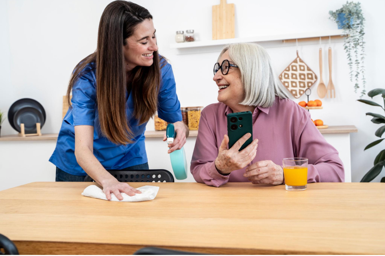 Two women smiling and talking at a table in a bright kitchen, one wiping the surface with a cloth.