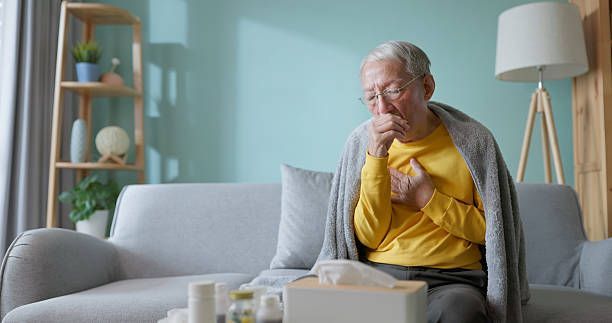 Older person coughing, clutching chest, seated on a sofa with tissues and medicine; indoor setting.