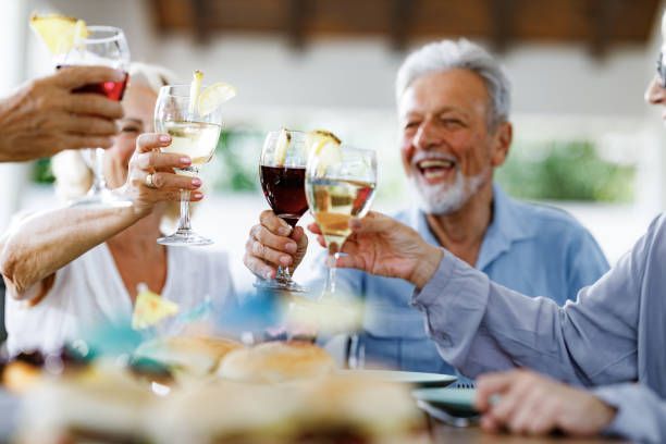 Elderly people clinking glasses of wine and cocktails, toasting at a sunny outdoor gathering.