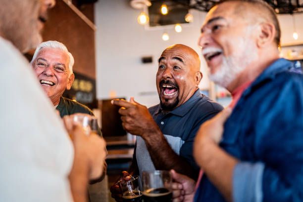 Four older men laughing and talking in a bar, holding alcoholic drinks.