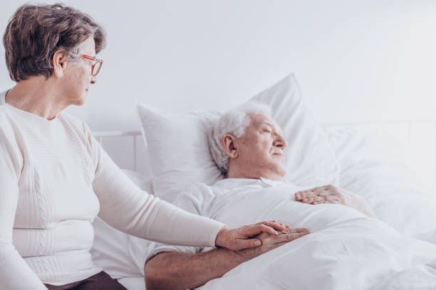 Elderly woman comforts an elderly man lying in bed, holding his hand