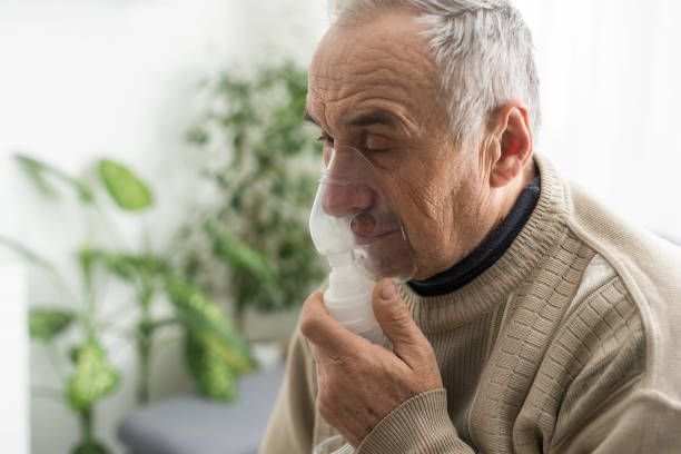 Man using a nebulizer, holding it up to his nose and mouth indoors. 