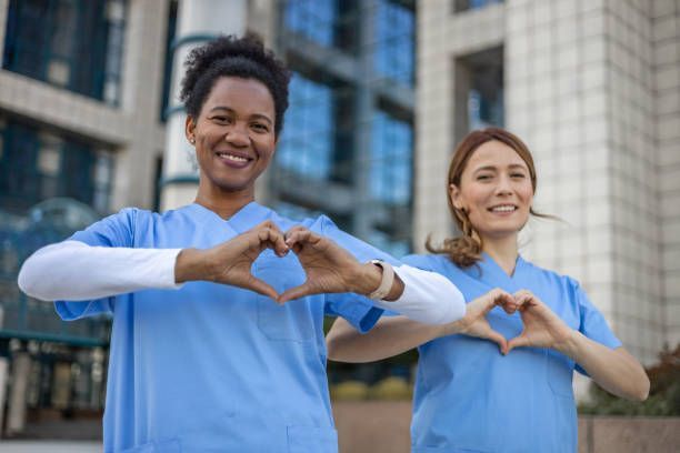 Two healthcare workers in blue scrubs making heart shapes with their hands, smiling outside a building.