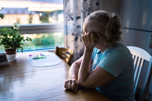 Isolated Woman with head in hand, seated at a table by a window; appears distressed.