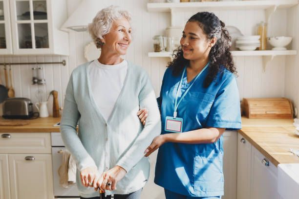 Elderly woman with cane and caregiver smiling in a kitchen. The caregiver has arm around the woman's back.
