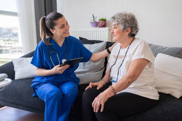 Nurse in blue scrubs, touching arm of senior seated on sofa, smiling and holding tablet.