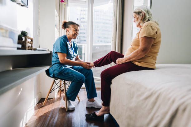 A healthcare worker examines a patient's leg while seated on a stool near a bed in a sunny room.