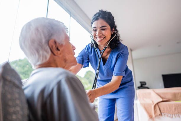 A smiling nurse in blue scrubs using a stethoscope on an elderly patient indoors.