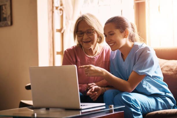 Young woman in blue scrubs points at laptop screen with older woman wearing glasses; indoor.