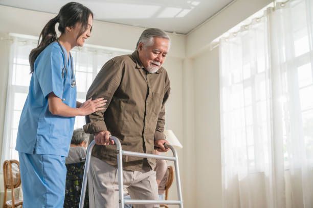Woman in blue scrubs assists older person using a walker in a light-filled room.