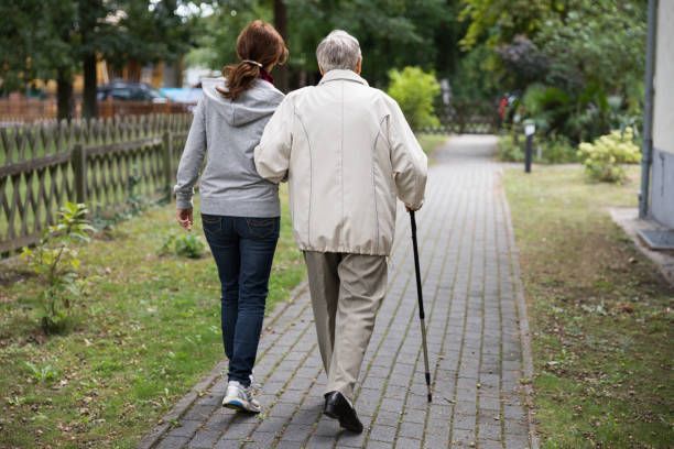 Woman assisting an elderly man with a cane walk on a brick path outside.