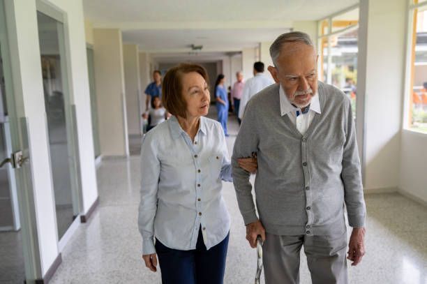 An older woman helps an older man walk down a bright hospital hallway, holding his arm.