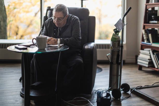 Man in chair, oxygen tank nearby, reading with coffee. Indoor, window light.