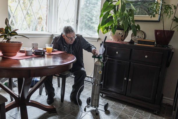 Man with oxygen tank at a dining table. He's adjusting the tank in a room with plants, a window, and cabinet.