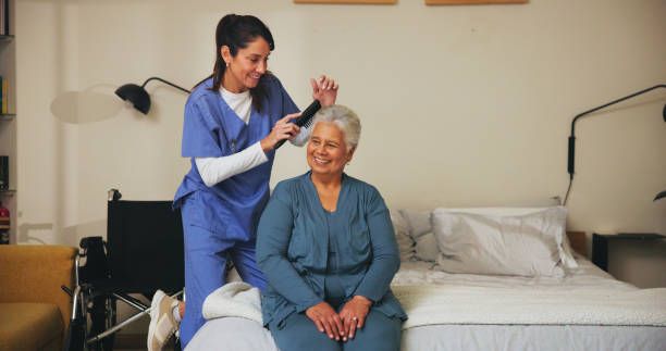 A smiling caregiver brushes an elderly woman’s hair while she sits on a bed; a wheelchair is nearby.