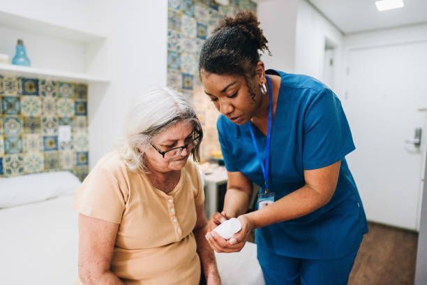 A home health aide shows medication to an older woman indoors; both are looking at the pills.