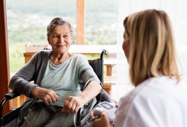 Elderly woman in wheelchair smiles at a nurse taking her blood pressure indoors near a window.