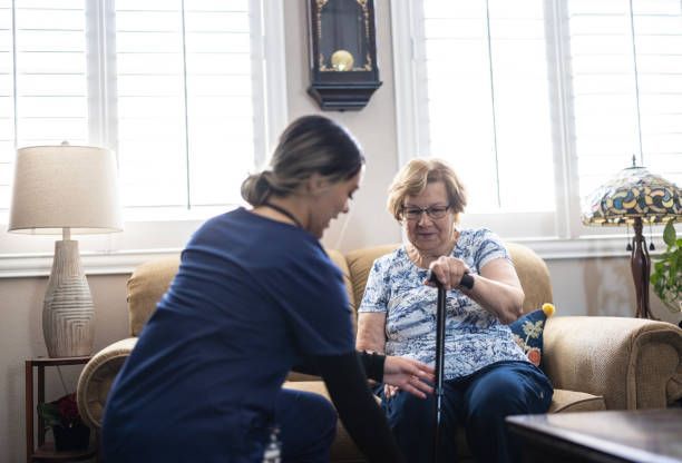 Caregiver helping an elderly woman with a cane to stand in a living room.