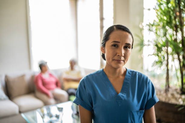 A healthcare worker in blue scrubs smiles in a care facility. Two elderly patients sit on a couch in the background.