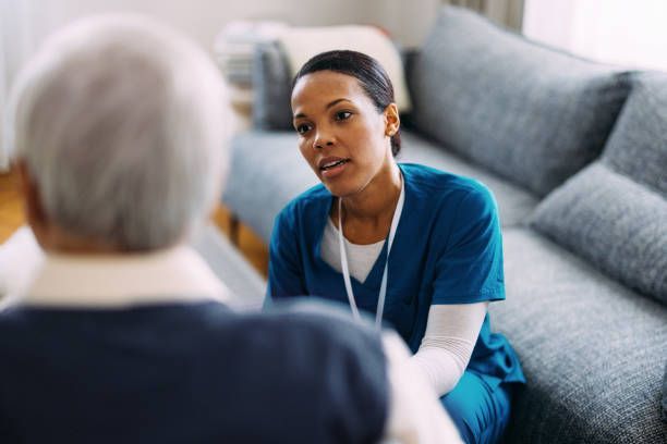 Caregiver in blue scrubs talking to a patient on a couch indoors.