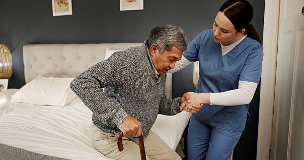 Caregiver assisting an older person to stand up from a bed; bedroom setting.