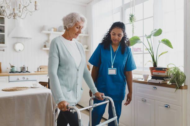 Woman with walker, assisted by a healthcare worker, inside a bright kitchen.