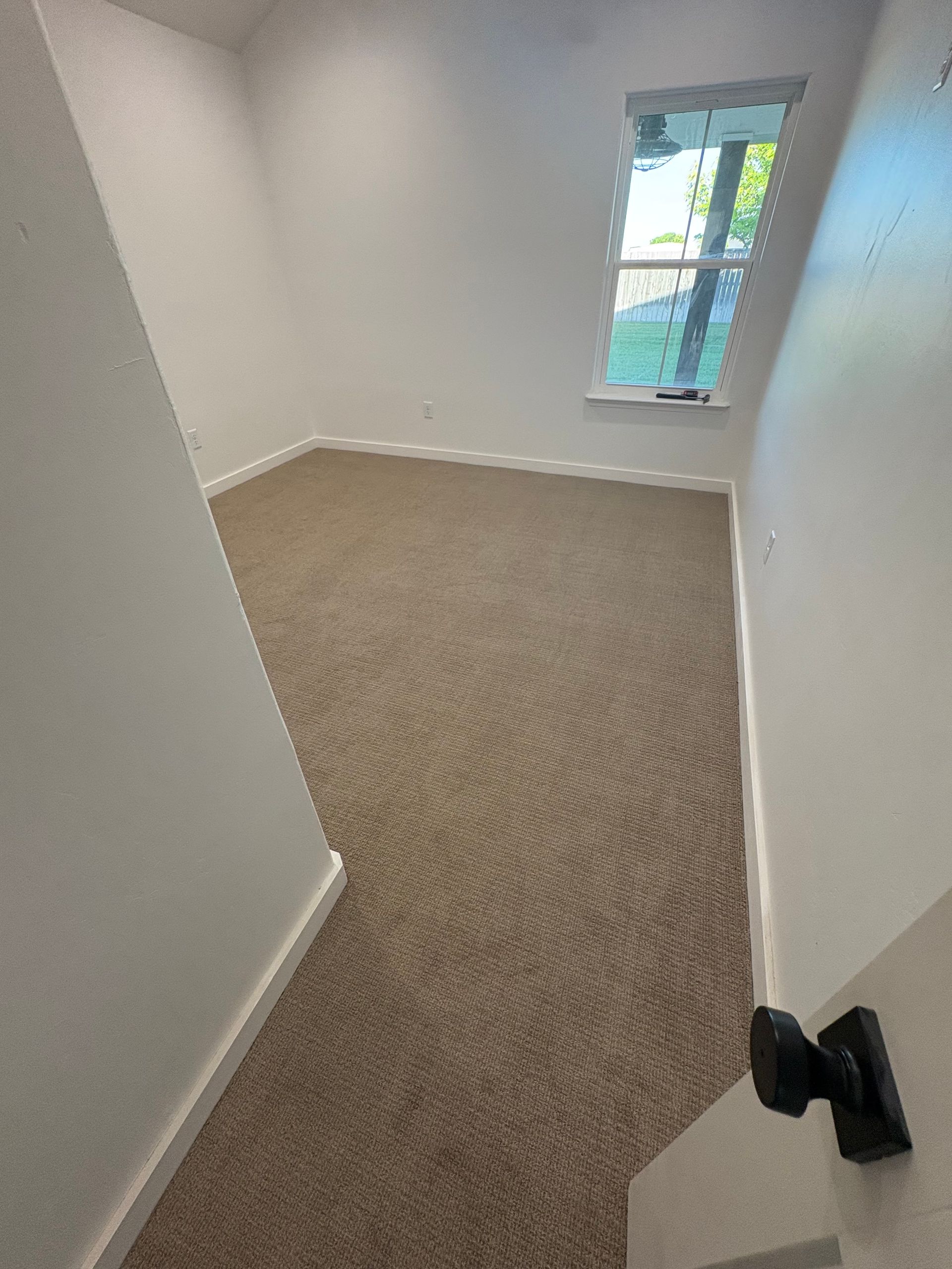 Empty living room with fireplace, kitchen, large windows, light gray walls, dark brown island, and gray carpet. Empty living room with fireplace, kitchen, large windows, light gray walls, dark brown island, and gray carpet.