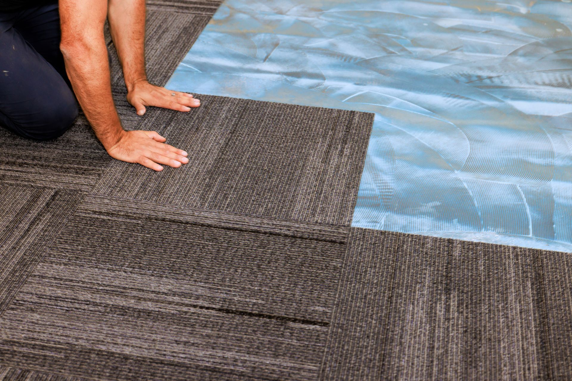 Person installing a gray carpet tile over blue adhesive film on an existing carpeted floor.