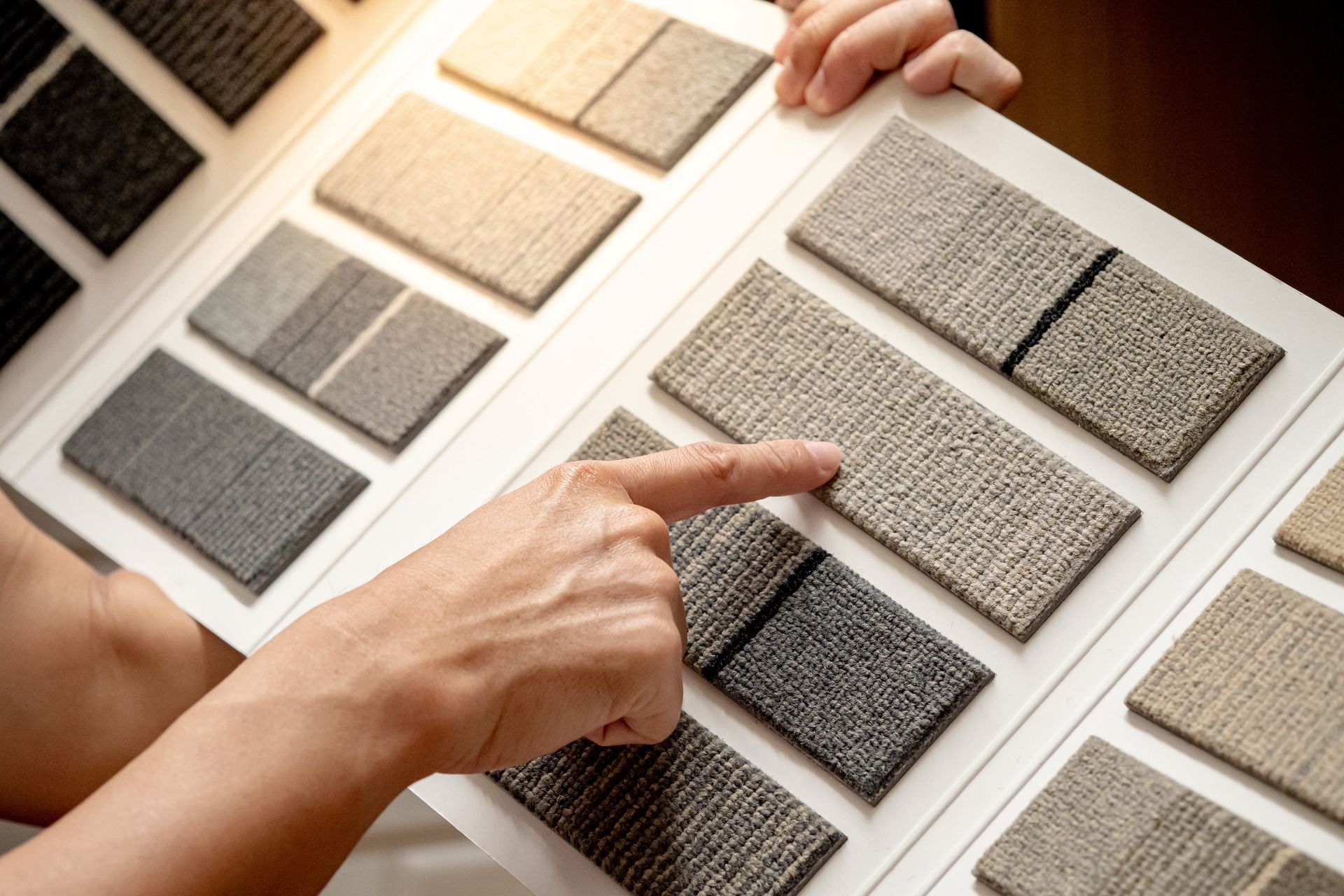 Hand pointing at a light gray carpet sample on a display board with other carpet swatches.