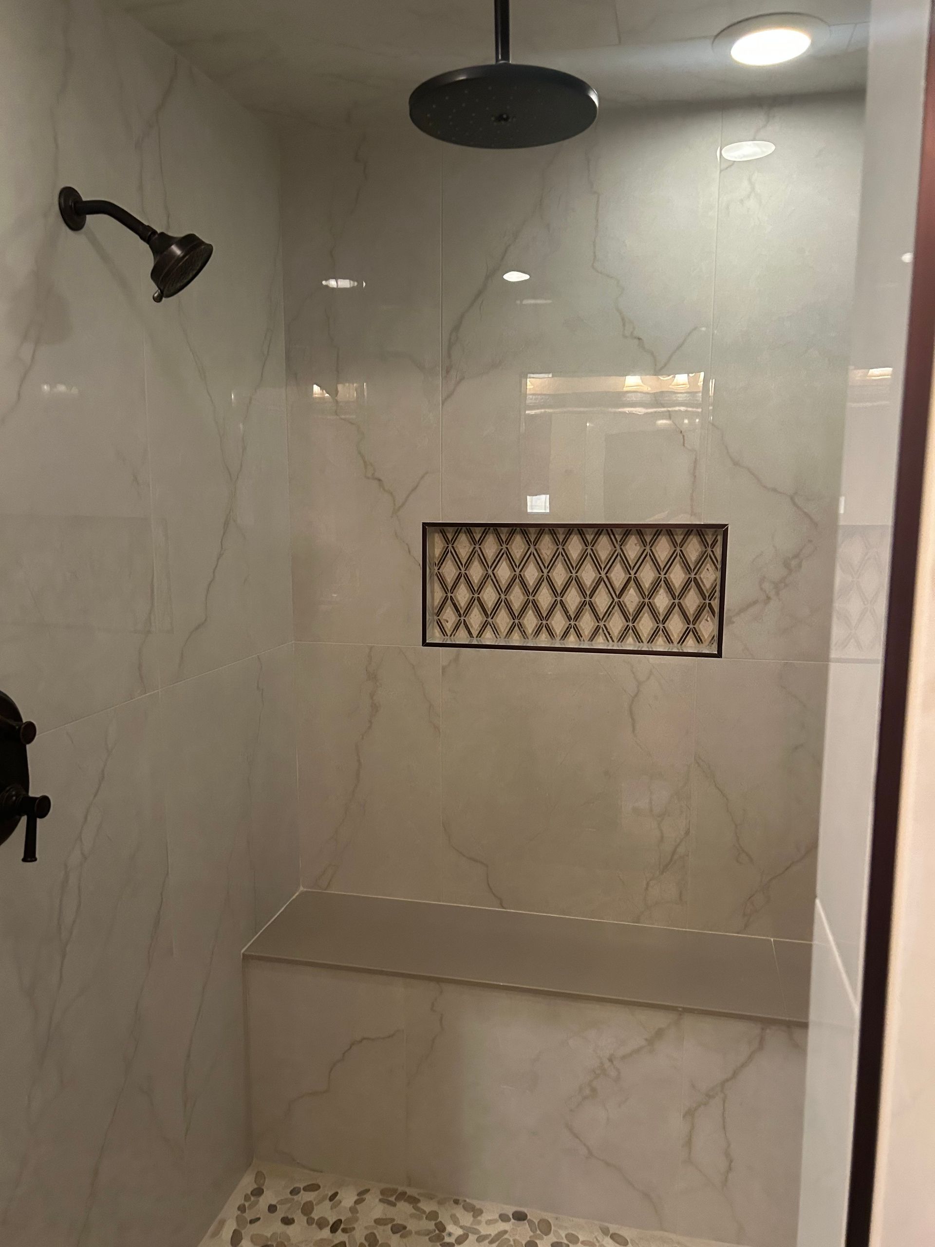 Shower with marble-look tile, dark fixtures, a built-in bench, and decorative niche.