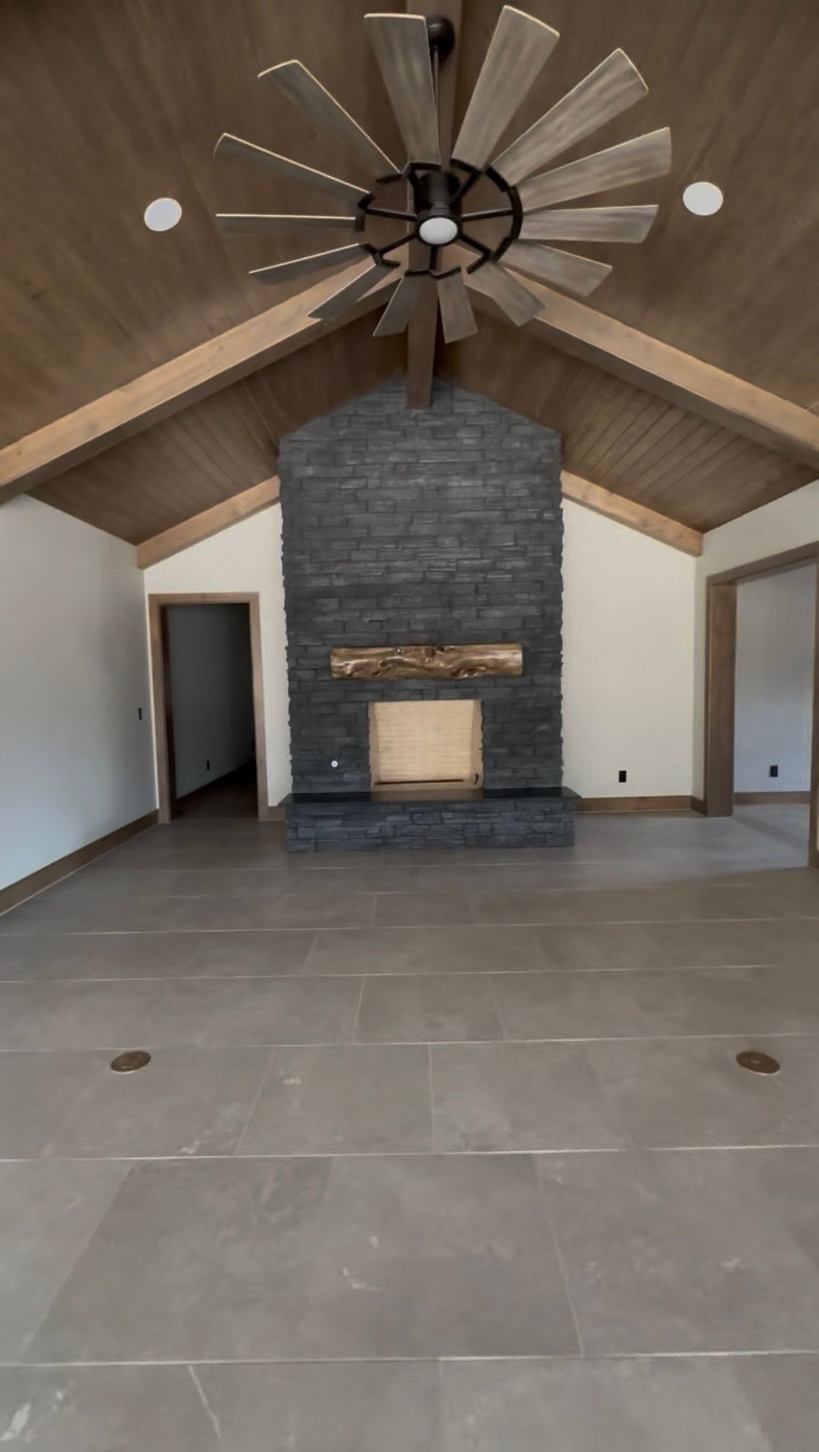 Spacious room with fireplace, wood ceiling, and ceiling fan. Gray tiled floor, neutral walls, and two doorways.