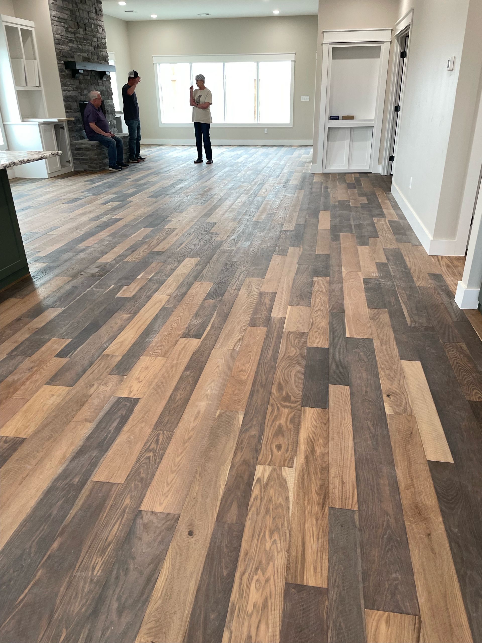 Wooden floor with brown and gray planks, with people standing in the background near a window and fireplace.