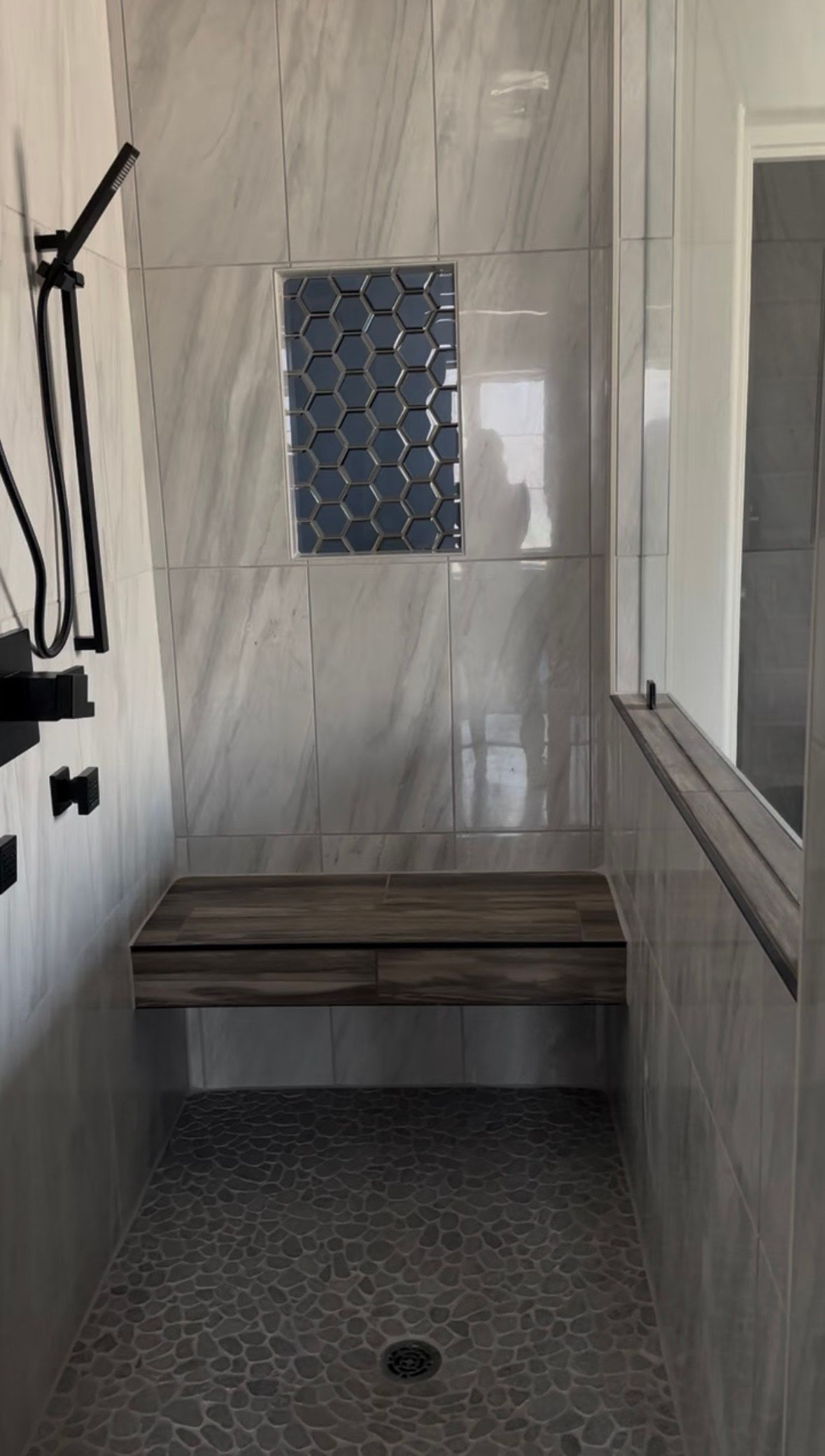 A modern shower with gray marbled tile walls, a built-in wooden bench, and black shower fixtures.