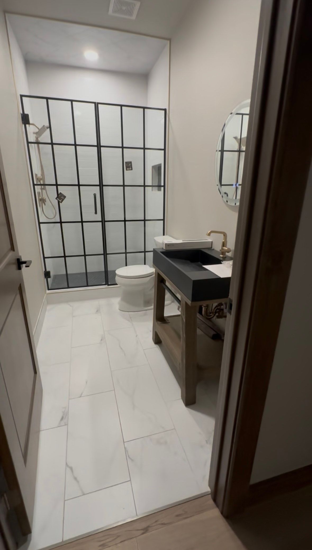 Bathroom with a black-framed shower, toilet, and vanity with a circular mirror. White tile floor.